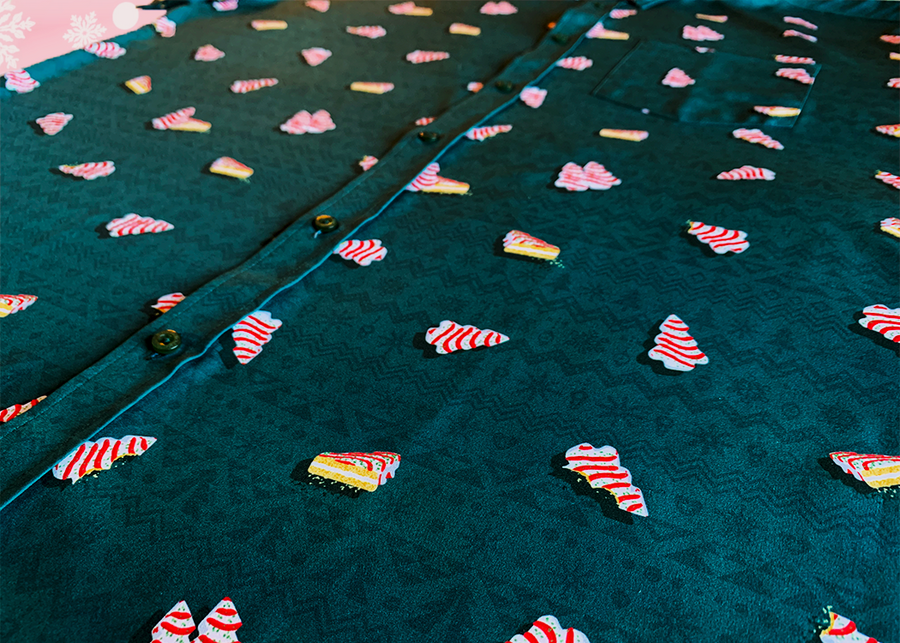 Angled view of the 7-Strong "Oh, Christmas Treat" shirt - a green, christmas sweater-like background with columns of various white Christmas tree shaped cakes with red garland, some whole, some bitten. The shirt is displayed against a red, snowing background. 