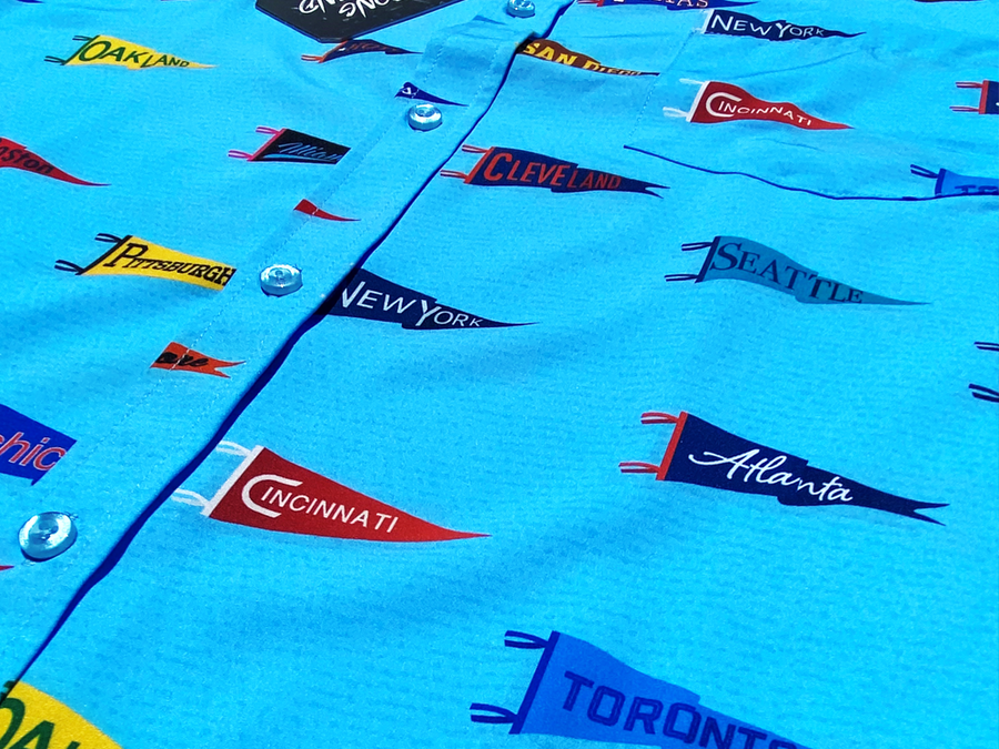 Close up of midsection of the 7-Strong "Pennant to Win It" shirt, which is sky blue with texture, featuring various triangular flags featuring baseball's prominent cities patterned throughout. The shirt is featured against abackground is sunset at a major league stadium.