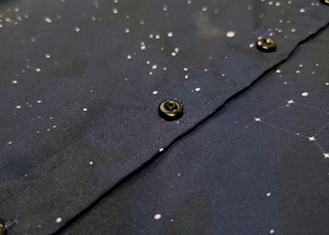 Midsection view of the short-sleeve Stargazer button down shirt. A deep navy blue shirt with constellation star patterns throughout. The shirt is displayed against a night sky full of stars.
