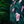 Load image into Gallery viewer, Side of Mid drift button view of the Youth 7-Strong &quot;Tropic Like It&#39;s Hot&quot; button down shirt - a black shirt with distinctive green palm trees and pink, white, and purple flowers peering out from behind the palms. Shirt is shown on a tropical rainforest background.
