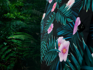 Side of Mid drift button view of the Youth 7-Strong "Tropic Like It's Hot" button down shirt - a black shirt with distinctive green palm trees and pink, white, and purple flowers peering out from behind the palms. Shirt is shown on a tropical rainforest background.
