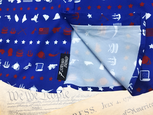 A close-up representation of 7-Strong's 1776 button down shirt, in the sweep tag region, against a Declaration of Independence background. Shirt is royal blue with stripes of various Americana icons, including the original 13 colonies and revolutionary war items.