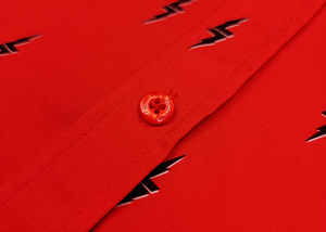 Close up mid button view of the 7-Strong Red 7-Bolt short sleeve shirt, featuring black 7-bolts with a white drop shadow interspersed on a red background. The shirt is featured on a red lightning storm backdrop.