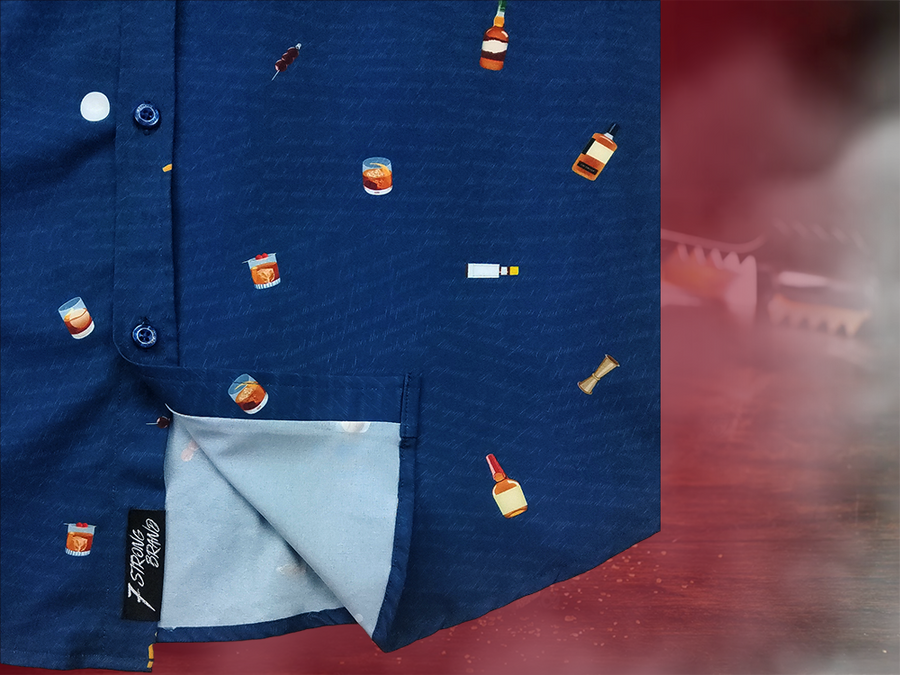 Bottom corner, sweep tag layout of "Call Me Old Fashioned" adult short sleeve button-down. Features elements of an Old Fashioned cocktail (bottle of bourbon, glasses, oranges, stirrers, and ice) against a blue background which faintly spells out an Old Fashioned recipe. Shirt is on a background featuring a finished Old Fashioned.