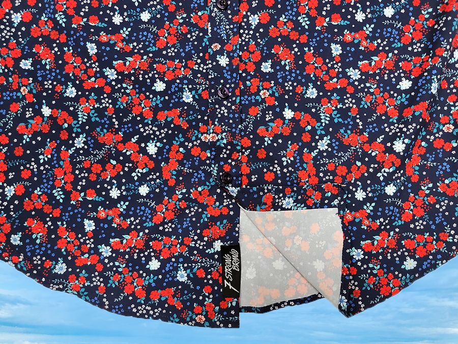 Bottom half sweep tag view of the 7-Strong "Aye Poppy" button down, featuring an array of red poppys with white sprigs on a deep navy blue shirt. The shirt is displayed against a partly cloudy sky.