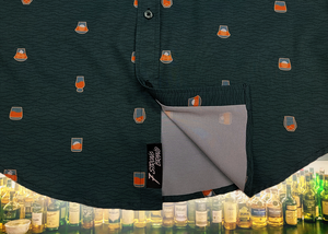 Bottom sweep tag view of the 7-Strong "Straight Sippin'" Short Sleeve button down. The shirt is a deep green with black ripples as its background, while the foreground has various drinking glasses with various levels of whiskey, some with ice, others without. The shirt is shown against a background of various whiskies and bourbons on a shelf.
