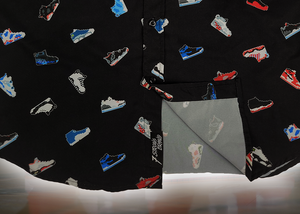 Bottom sweep tag view of the adult short sleeve version of "Got 'Em!" The button down features various athletic fashion sneakers in an 8-bit format arranged all over a black shirt. The shirt is featured against a background of a basketball player on the court, noticeable sneakers being worn