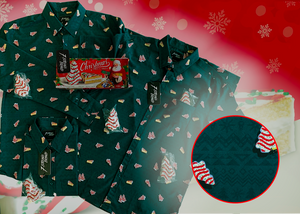 Full view of the long-sleeve, adult and youth short-sleeve versions of the 7-Strong "Oh, Christmas Treat" shirt overlapping each other with various cakes laid on top- a green, christmas sweater-like background with columns of various white Christmas tree shaped cakes with red garland, some whole, some bitten. The shirt is displayed against a red, snowing background with a similar parted cake. Bottom right features a detail circle showcasing the shirt's design. 