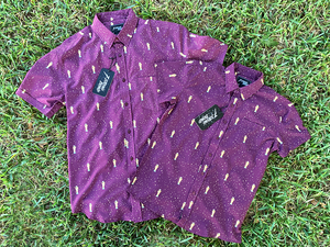 Full view of the Cup of Life adult and youth button ups, overlapping one another, a off-maroon colored shirt decorated throughout with falling gold confetti and depictions of the World Cup statue. The shirt is laid out on a grass pitch.