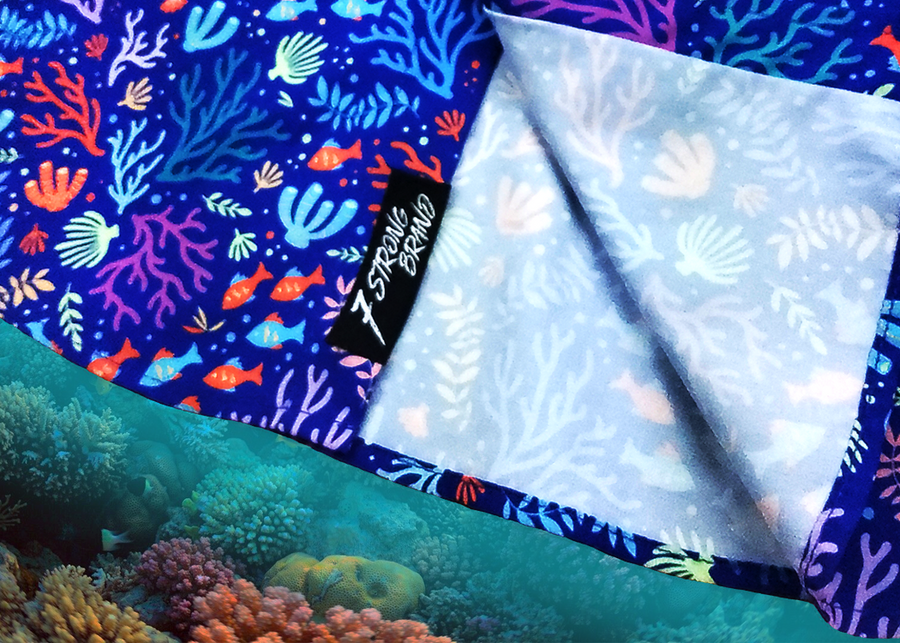Bottom sweep tag area shot of the 7-Strong "Coral of the Story" adult button down shirt. Color of the shirt is blue with vibrant purple, teal, orange sea life imagery - particularly fish and coral reef. The shirt is shown against an underwater background showcasing coral.