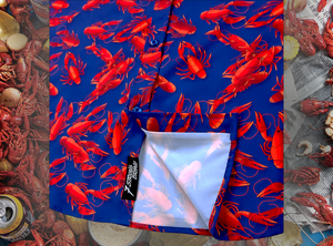Close up view of the 7-Strong Dat Boil shirt sweep tag, along with design of shirt in deep navy blue with red crawfish patterned throughout overlapping one another. The shirt itself sits on a background image of items from a Crawfish Boil such as crawfish, seasoning, corn, and potatoes. 