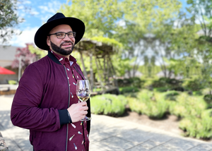 Medium close up of a model smiling towards camera wearing the 7-Strong "Pour Decisions" button down, layered with a maroon bomber jacket and accentuated with a wide brim black fedora - holding a glass of wine in hand at a vineyard. 