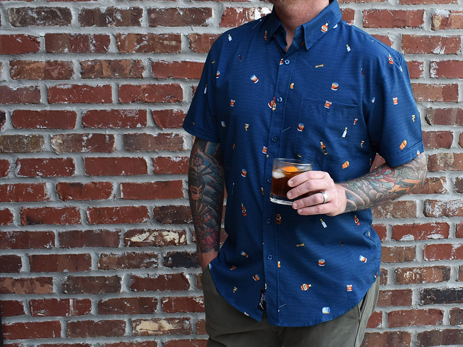 Adult male model, holding an Old Fashioned and wearing "Call Me Old Fashioned" adult short sleeve button-down. Features elements of an Old Fashioned cocktail (bottle of bourbon, glasses, oranges, stirrers, and ice) against a blue background which faintly spells out an Old Fashioned recipe. Model is against a brick wall. 