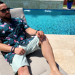 Load image into Gallery viewer, Male adult model sitting in a beach recliner by the pool wearing the 7-Strong &quot;Tropic Like It&#39;s Hot&quot; adult button up. Shirt is black with detailed green palms, matches with white, pink, and purple flowers peering throughout the shirt - paired with light mint shorts. 
