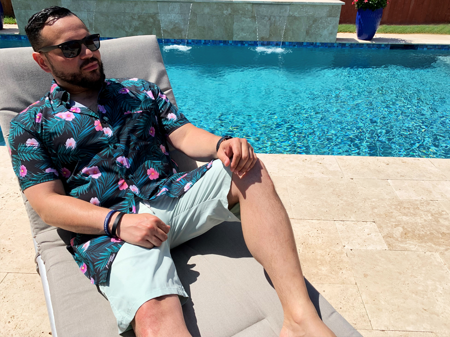Male adult model sitting in a beach recliner by the pool wearing the 7-Strong "Tropic Like It's Hot" adult button up. Shirt is black with detailed green palms, matches with white, pink, and purple flowers peering throughout the shirt - paired with light mint shorts. 