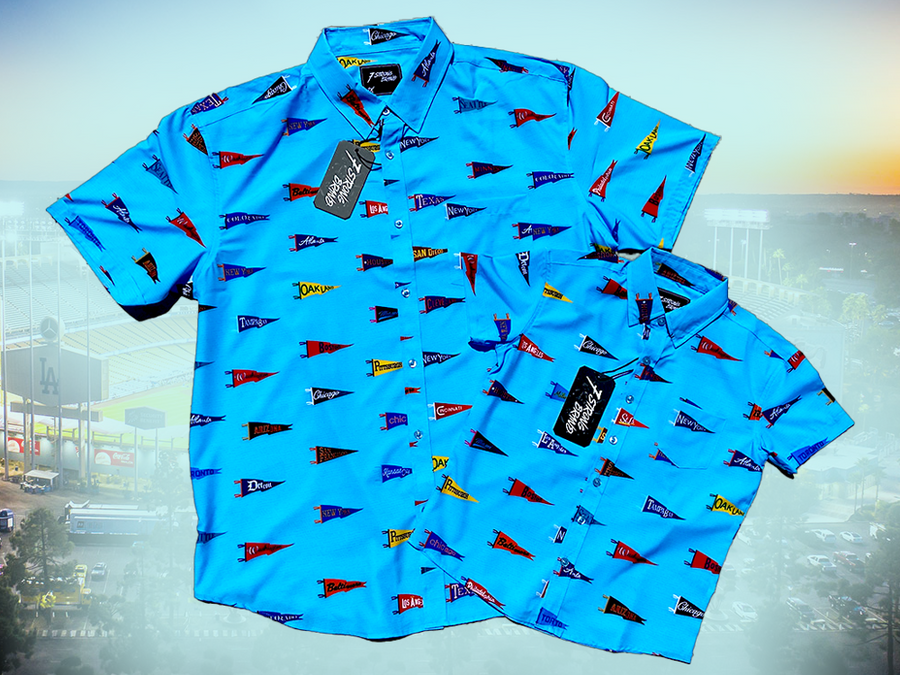 Full view of both the adult and youth 7-Strong "Pennant to Win It" shirt, which is sky blue with texture, featuring various triangular flags featuring baseball's prominent cities patterned throughout. The shirt is featured against abackground is sunset at a major league stadium.