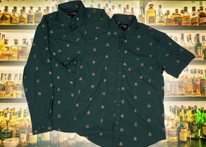 Full display view of the 7-Strong "Straight Sippin'" Short Sleeve and long-sleeve button downs, overlapping each other. The shirt is a deep green with black ripples as its background, while the foreground has various drinking glasses with various levels of whiskey, some with ice, others without. The shirt is shown against a background of various whiskies and bourbons on a shelf.