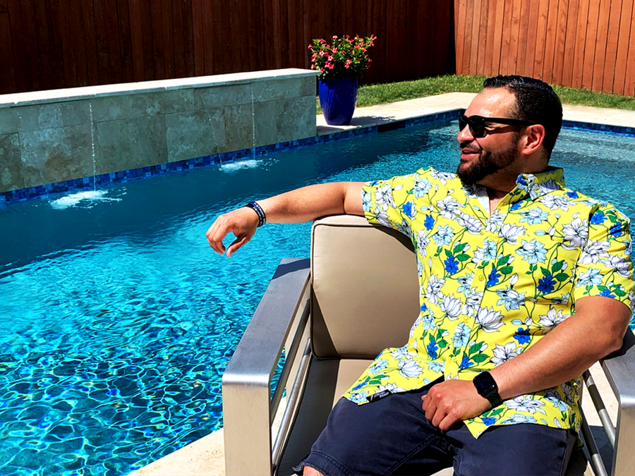 Male model wearing the 7-Strong "Late Bloomer" adult button down - a yellow background shirt with white and blue flowers patterned all over it. Model is sitting by the pool in a chair, looking over the water, in sunglasses, smiling.  