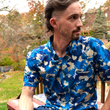 Load image into Gallery viewer, Male model of medium build, sitting in nature looking off sporting the 7-Strong &quot;Service Stars&quot; Adult button-up, featuring blue and creme colored camouflage with white weathered stars throughout. 

