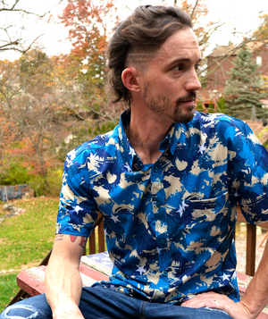Male model of medium build, sitting in nature looking off sporting the 7-Strong "Service Stars" Adult button-up, featuring blue and creme colored camouflage with white weathered stars throughout. 