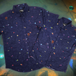 Load image into Gallery viewer, Full view shot of both adult and youth 7-Strong &quot;Mine Train&quot; button downs overlapping each other. Shirt is a deep purple with a rock quarry depicting ghosted design showcasing various colors of gemstones and mine train carts throughout. The shirt itself is displayed against a background of a mine shaft and various gems found in buckets. 
