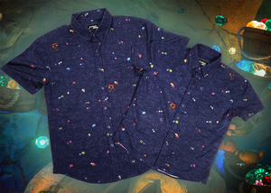 Full view shot of both adult and youth 7-Strong "Mine Train" button downs overlapping each other. Shirt is a deep purple with a rock quarry depicting ghosted design showcasing various colors of gemstones and mine train carts throughout. The shirt itself is displayed against a background of a mine shaft and various gems found in buckets. 