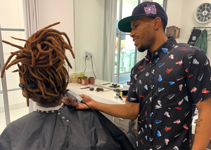 Male model wearing he adult short sleeve version of "Got 'Em!" The button down features various athletic fashion sneakers in an 8-bit format arranged all over a black shirt.  Model is a barber shaping up the hairstyle of a patron.