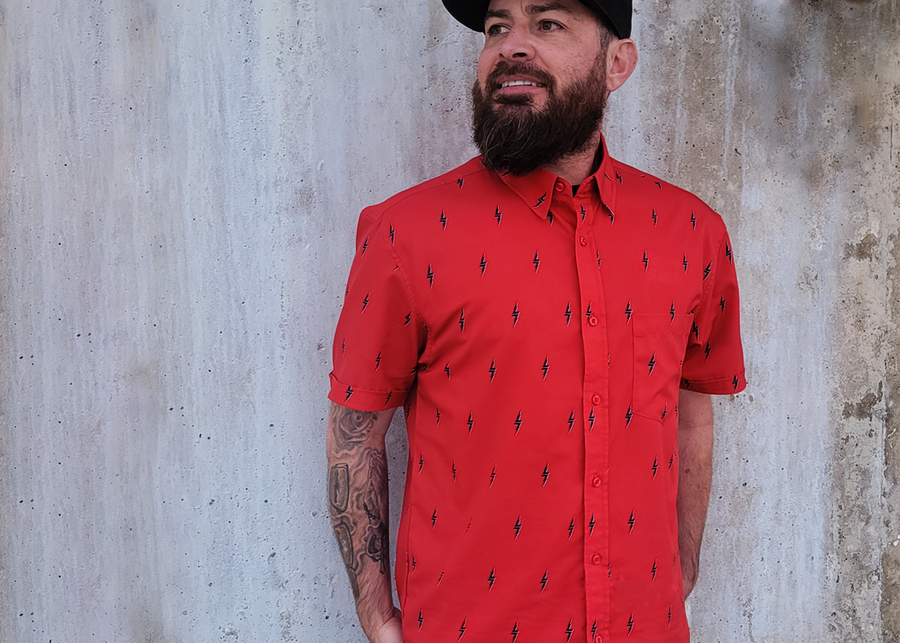 Medium shot of a model against a wall wearing the  7-Strong Red 7-Bolt short sleeve shirt, featuring black 7-bolts with a white drop shadow interspersed on a red background.
