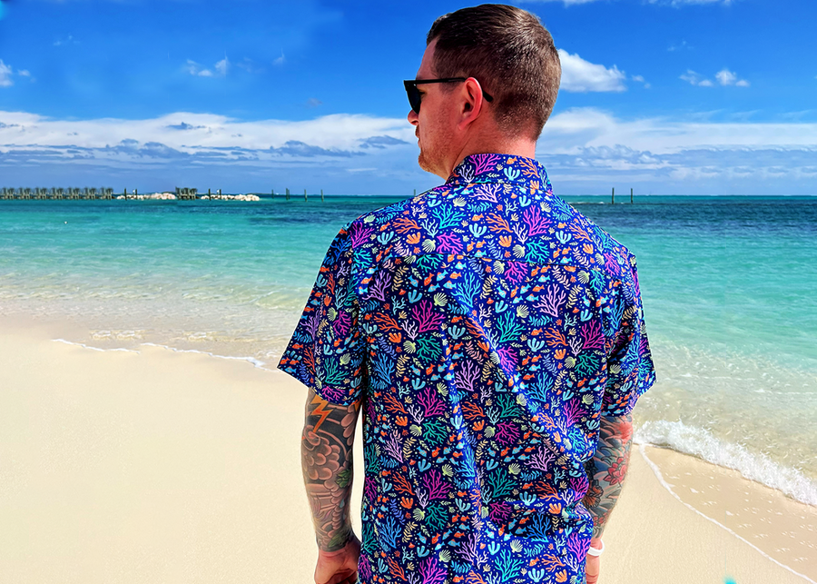 Medium close up, behind the back view of a male model of medium build wearing the 7-Strong "Coral of the Story" adult button down shirt. Color of the shirt is blue with vibrant purple, teal, orange sea life imagery - particularly fish and coral reef. The model is standing on the beach looking out towards the water wearing the shirt. 