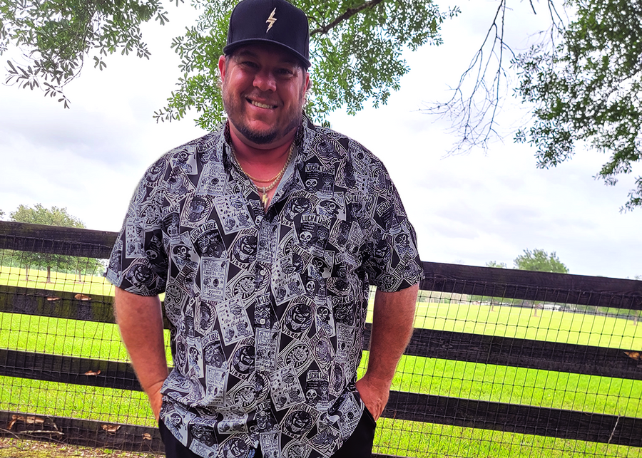 Male model wearing the adult 7-Strong "Cinco de Mayhem" button down shirt. Shirt features black and white posters featuring various created luchadors advertised for matches, all overlapping each other over a black shirt. Model is outside against a fence. 