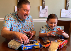 Father and son models wearing the 7-Strong "Cheat Day" adult button down featuring various snacks, treats, and fast food features on a light blue shirt. Both models are indulging in cookies and treats. 