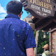 Load image into Gallery viewer, Medium shot of male model, back to camera, wearing 7-Strong Mine Train button down depicting various colored gemstones in a mine shaft of deep purple color. Model is facing the entrance to Mine Train attraction. 

