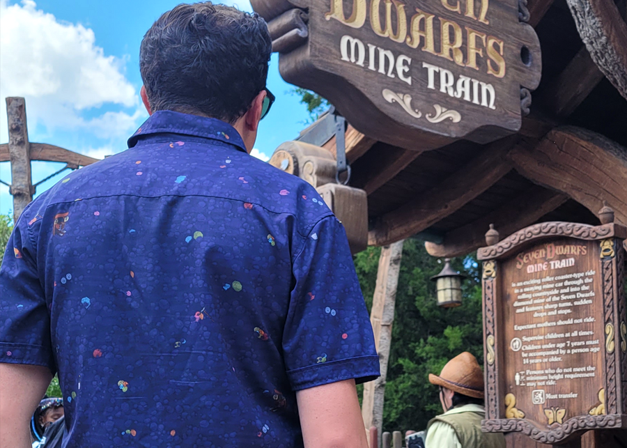 Medium shot of male model, back to camera, wearing 7-Strong Mine Train button down depicting various colored gemstones in a mine shaft of deep purple color. Model is facing the entrance to Mine Train attraction. 