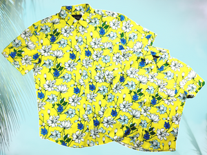 Full view of both the 7-Strong "Late Bloomer" adult and youth button downs overlapping each other - a yellow background shirt with white and blue flowers patterned all over it. Shirt is against a blue tropical sky with palm trees in the distance. 