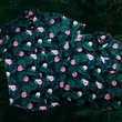 Load image into Gallery viewer, Full view of both the Adult and Youth 7-Strong &quot;Tropic Like It&#39;s Hot&quot; button down shirts overlapping one another - a black shirt with distinctive green palm trees and pink, white, and purple flowers peering out from behind the palms. Shirt is shown on a tropical rainforest background.
