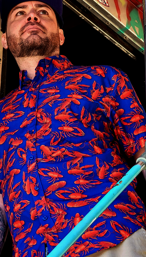Medium shot of adult male model of medium build wearing the 7-Strong Dat Boil shirt in deep navy blue with red crawfish patterned throughout overlapping one another. 