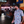 Load image into Gallery viewer, Medium shot of adult male in the 7-Strong Block Party adult button down, featuring various colored blocks of different arrangements inspired by a classic video game, cascading down the shirt against a grayish background featuring shadows of the same shapes. Model is posed in the lobby of an arcade with &quot;7-STRONG BRAND&quot; on its marquee.  
