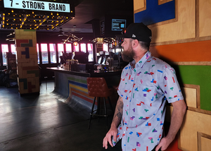 Medium shot of adult male in the 7-Strong Block Party adult button down, featuring various colored blocks of different arrangements inspired by a classic video game, cascading down the shirt against a grayish background featuring shadows of the same shapes. Model is posed in the lobby of an arcade with "7-STRONG BRAND" on its marquee.  