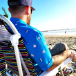 Load image into Gallery viewer, Behind the back shot of a male model sitting on the beach with the &quot;When Pigs Fly&quot; shirt being worn, sitting on a beach chair and looking out towards water. 
