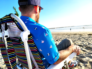 Behind the back shot of a male model sitting on the beach with the "When Pigs Fly" shirt being worn, sitting on a beach chair and looking out towards water. 