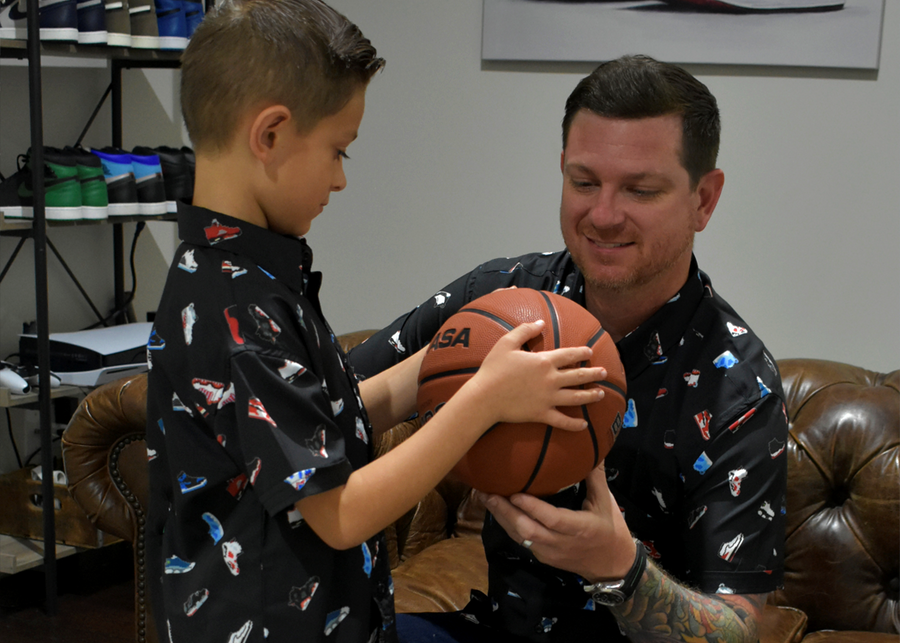 Father and son model shot, both wearing their version of the short sleeve "Got 'Em!" The button down features various athletic fashion sneakers in an 8-bit format arranged all over a black shirt. Both are admiring a basketball held by the child. 