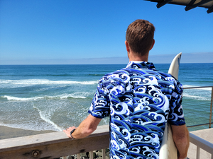 Male model, back to camera holding a surfboard overlooking the ocean while wearing the 7-Strong "Let 'Er Riptipe" Adult button down shirt, featuring an array of indigo and purple waves with whitecaps all over. 