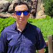 Load image into Gallery viewer, Medium shot of a average male model in sunglasses wearing the 7-Strong Mine Train shirt depicting various gemstones on a rock quarry background in a deep purple. He stands in front of the Mine Train attraction as its in movement. 
