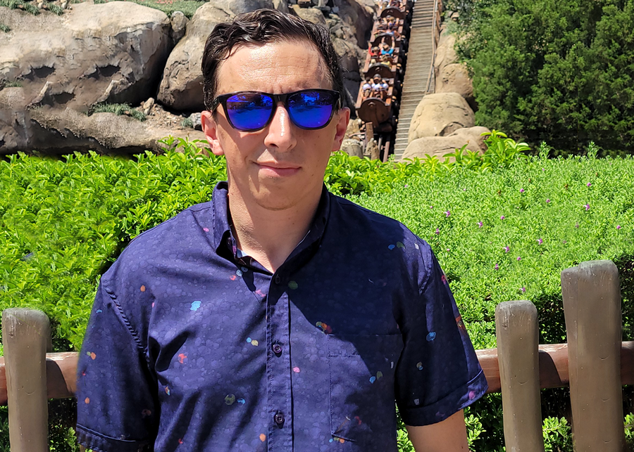 Medium shot of a average male model in sunglasses wearing the 7-Strong Mine Train shirt depicting various gemstones on a rock quarry background in a deep purple. He stands in front of the Mine Train attraction as its in movement. 