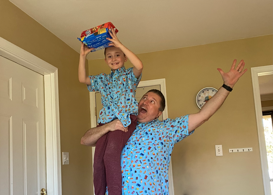Father and son model sporting the 7-Strong "Cheat Day" Shirts. Father has son on his shoulder in the air while the son holds cookie packages in his hand. 