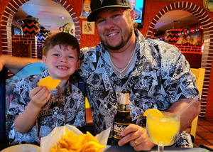 Father and son at a Mexican restaurant smiling at camera wearing the youth and adult adult 7-Strong "Cinco de Mayhem" button down shirt. Shirt features black and white posters featuring various created luchadors advertised for matches, all overlapping each other over a black shirt. 