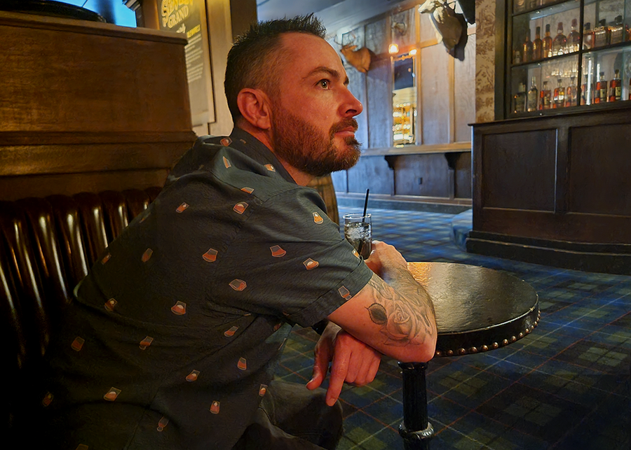 Male model wearing the 7-Strong "Straight Sippin'" Short Sleeve button down. The shirt is a deep green with black ripples as its background, while the foreground has various drinking glasses with various levels of whiskey, some with ice, others without. The model is sitting at a table with a drink inside a bourbon bar. 