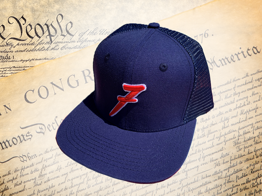Full, slight overhead shot of the 7-Series trucker hat, showcasing its all navy blue look, back mesh panel, red puffed embroidered 7 with white outline, against a background of the Declaration of Independence.