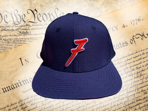Full, head-on shot of the 7-Series trucker hat, showcasing its all navy blue look, solid front panels, red puffed embroidered 7 with white outline, against a background of the Declaration of Independence.