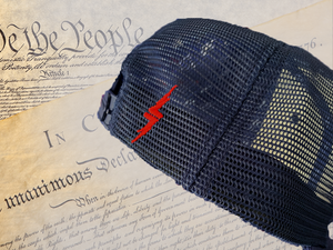 Back mesh panels view of 7-Series Red, White, and Blue trucker hat, featuring a red 7-Bolt logo on the side beside the snapback. Hat is shown against backdrop of the Declaration of Independence. 